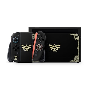 Puede incluir: Una consola de videojuegos negra y un mando a juego con emblemas dorados de la Trifuerza y patrones intrincados inspirados en Hylia. La base de la consola y el mando muestran gráficos dorados estilizados, incluyendo un icono de espada.