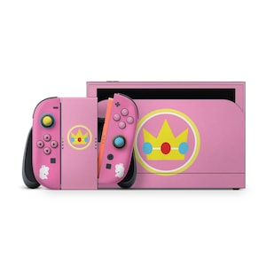 May include: A pink video game console dock and two matching controllers. The dock features a large yellow crown emblem with red and blue circles. The controllers have pink shells, yellow and blue thumbsticks, and small white crown icons.