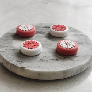 May include: Four decorative, round objects with floral designs rest on a gray marble tray. The objects have a white base and a red top, with a white or red flower design. The tray is circular with a smooth, polished surface.