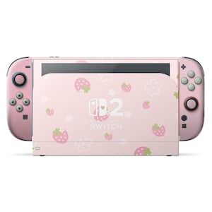 Peut inclure: Une console Nintendo Switch rose avec des manettes assorties. La console présente un fond rose clair avec un motif de fraises et de fleurs. Le logo Nintendo Switch est centré sur l'écran.