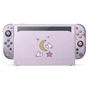 May include: A lavender Nintendo Switch console with matching controllers. The console and controllers are decorated with cartoon bunnies, crescent moons, and stars. The design is a cute and whimsical theme.