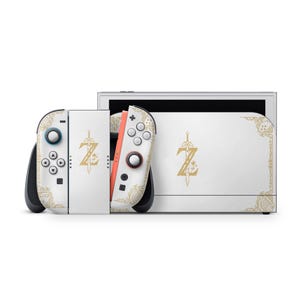 May include: A white video game console and two controllers featuring gold-colored decorative patterns and a central gold sword and Z emblem. The controllers have white buttons and joysticks, with one controller showing a red side edge.