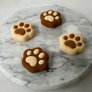 Peut inclure: Quatre décorations en forme de pattes, marron et crème. Les empreintes de pattes sont disposées sur une surface ronde en marbre. Ces articles sont probablement destinés à la décoration ou à l'artisanat.