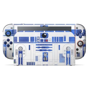 Puede incluir: Una consola Nintendo Switch con una piel personalizada que presenta un diseño azul y blanco que recuerda al personaje de Star Wars R2-D2. La piel cubre la consola y los mandos Joy-Con, con gráficos detallados.