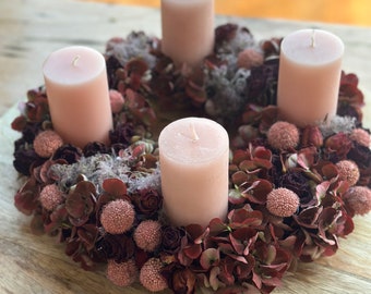 May include: A decorative candle wreath featuring four light pink pillar candles. The wreath is adorned with dried burgundy flowers, moss, and small, round, pink accents. The arrangement sits on a wooden surface, creating a warm, inviting aesthetic.