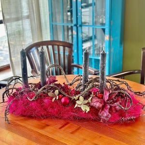 Könnte beinhalten: Festlicher Tischschmuck mit vier schwarzen Kerzen in einem leuchtend rosa Nest. Die Anordnung umfasst trockene Zweige, rote Beeren und florale Akzente, ideal für Feiertage.