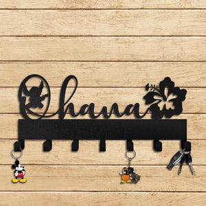 Peut inclure: Porte-clés en métal noir avec le mot "Ohana" et un motif de fleur d'hibiscus. Le porte-clés a 7 crochets et est monté sur un mur en bois. Il y a des clés et un porte-clés Mickey Mouse accrochés aux crochets.