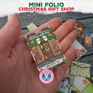 Puede incluir: Un mini folio de papel con una ilustración de una tienda de regalos navideños, con un toldo a rayas verdes y blancas, una puerta central con una corona y escaparates. El texto dice Christmas Gift Shop y Christmas Joy.