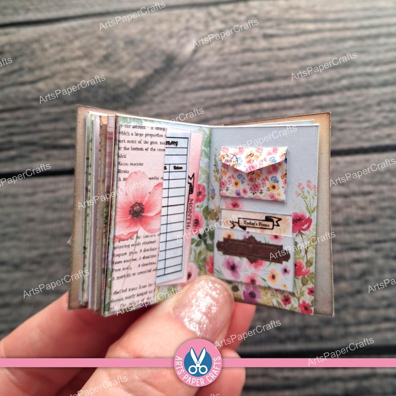 May include: A miniature open book featuring floral patterned pages, a printed grid chart, and two small attached envelopes. The left page displays a pink poppy illustration and text, while the right page includes a small floral envelope and a pocket.