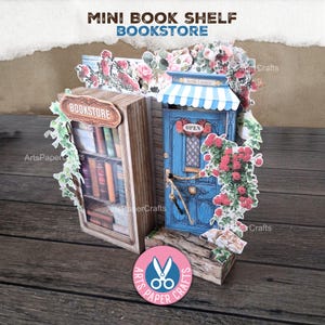 May include: A miniature papercraft bookstore featuring a bookshelf and a blue door. The bookshelf is filled with books, and the door has an "OPEN" sign. The scene is decorated with flowers and the text "MINI BOOK SHELF BOOKSTORE".