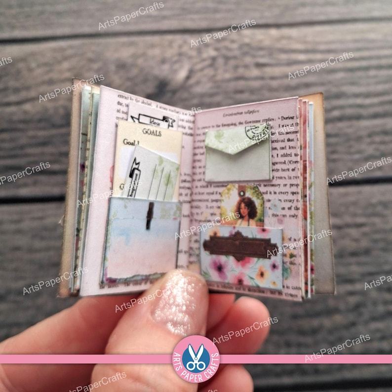 May include: A miniature open book featuring printed text, a small envelope, a card labelled Goals, and a floral pocket. The pages contain various paper inserts, including a small portrait and decorative elements, all presented in a compact format.