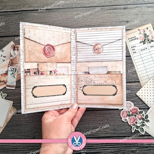 May include: Open journal with vintage-style pages, envelopes, and pockets. Features a rose-sealed envelope and a bird-sealed envelope. Includes decorative labels and floral accents. The journal is held open, showcasing its contents.