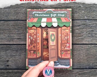 May include: A rectangular paper folio featuring a detailed illustration of a Christmas gift shop with a green and white striped awning, a partially open door, and windows displaying teddy bears, ornaments, and gifts. The text reads Christmas Gift Shop.