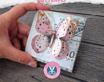 May include: A square tab folder featuring a three-dimensional butterfly with pink and white wings. The background displays a light blue vintage-style pattern with black printed text and numbers. The text TAB FOLDER BUTTERFLY is visible at the top.