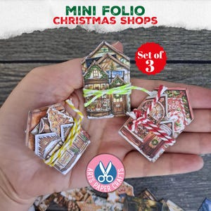 Puede incluir: Un conjunto de tres folios de papel en miniatura con ilustraciones de tiendas navideñas. Cada folio con forma de casa está atado con cordel decorativo amarillo, verde o rojo y blanco. Los diseños impresos muestran fachadas de tiendas.