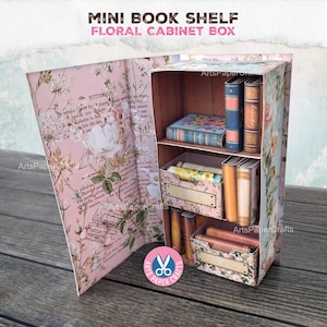 May include: A miniature floral cabinet box with a mini bookshelf design. The box is decorated with a pink floral pattern and contains small books and storage boxes. The text "MINI BOOK SHELF FLORAL CABINET BOX" is displayed at the top.