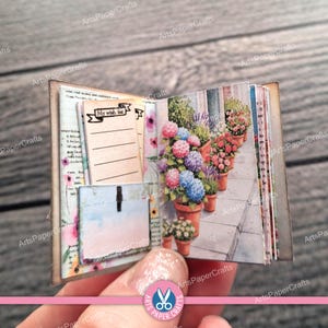 May include: A miniature open book featuring a left page with a My wish list notepad and a light blue pocket, and a right page displaying a printed illustration of potted pink and blue hydrangea flowers on a stone patio with a brick wall.