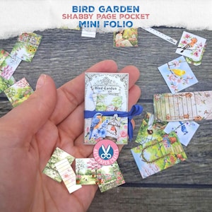 Op de afbeelding: Een miniatuur "Bird Garden" mini folio met een blauw lint, omringd door kleine, geïllustreerde kaarten. De folio heeft een blauwe vogel en kooi ontwerp. De tekst "Bird Garden Shabby Page Pocket Mini Folio" is zichtbaar.