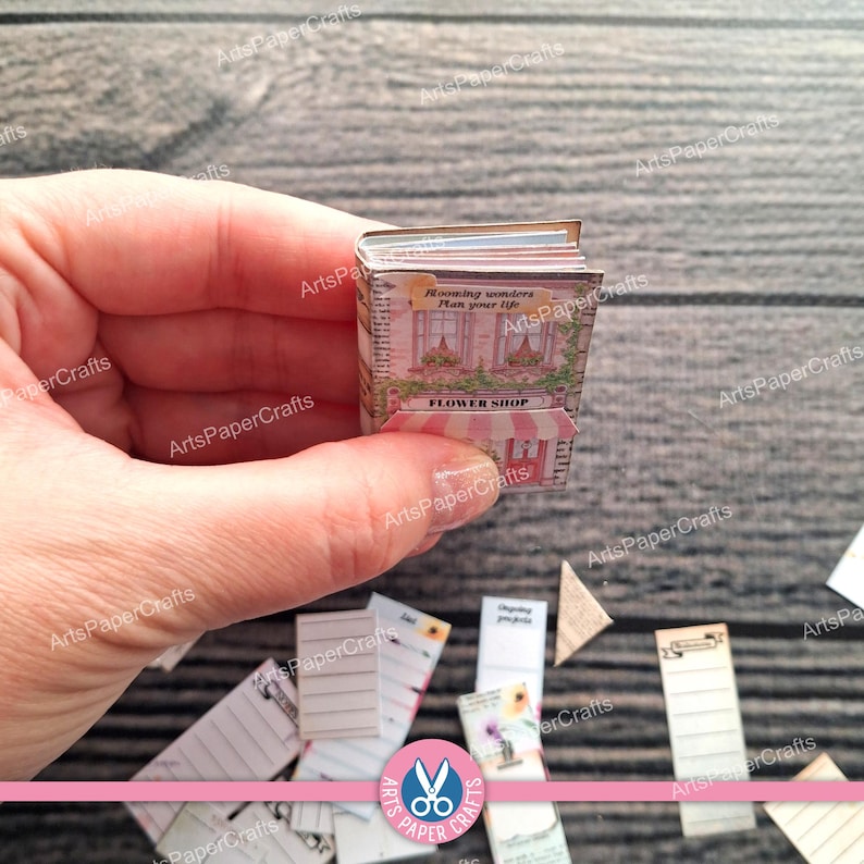 May include: A miniature book with a pink and white facade illustration on the cover. The cover features the text "Blooming wonders Plan your life" and "FLOWER SHOP" in black lettering. The book has visible white pages and a striped awning detail.