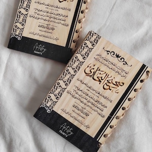 Puede incluir: Dos cuadernos de madera con bordes negros y diseños intrincados. Las portadas presentan texto árabe en un estilo tradicional. El texto dice "Sahih al-Bukhari", que es el nombre de una famosa colección de hadices, o dichos del Profeta Mahoma. Los cuadernos están encuadernados con una cuerda marrón.