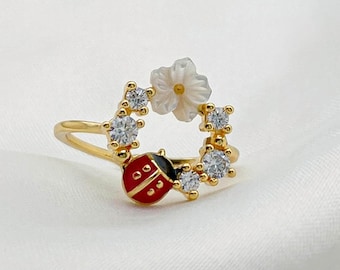 May include: A gold-toned ring featuring a ladybug and floral design. The ring has a red ladybug with black spots, a white flower, and clear gemstones. The ring is set against a white, textured background.