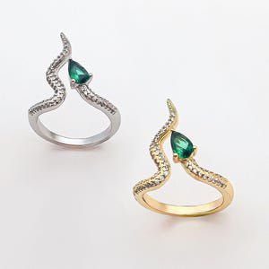 Puede incluir: Dos anillos, uno plateado y otro dorado, sobre un fondo blanco. Cada anillo presenta un diseño fluido y abstracto con una gema verde en forma de lágrima. El anillo plateado está adornado con pequeñas piedras transparentes, mientras que el dorado tiene piedras similares.