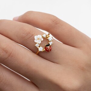 May include: A gold-colored ring featuring a wreath design with a white flower, clear gemstones, and a red and black ladybug. The ring is worn on a finger, showcasing the intricate details of the jewelry.