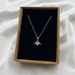 Puede incluir: Un collar de cadena dorada con un colgante de coraz&oacute;n rosa con peque&ntilde;as alas doradas. El collar se muestra en una caja marr&oacute;n con un interior de terciopelo negro.
