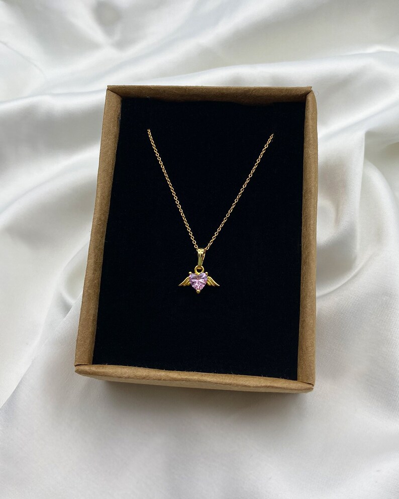 Puede incluir: Un collar de cadena dorada con un colgante de coraz&oacute;n rosa con peque&ntilde;as alas doradas. El collar se muestra en una caja marr&oacute;n con un interior de terciopelo negro.