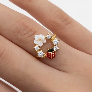 May include: A gold-colored ring featuring a floral design with a white flower and a red ladybug. The ring is adorned with clear, round gemstones. The ring is a piece of jewelry.