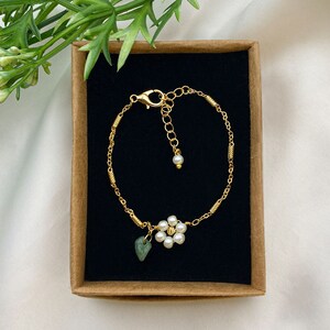 Pu&ograve; includere: Un braccialetto a catena dorata con un delicato ciondolo a forma di fiore realizzato con perle bianche e una pietra verde. Il braccialetto &egrave; esposto in una scatola marrone con un rivestimento in velluto nero.