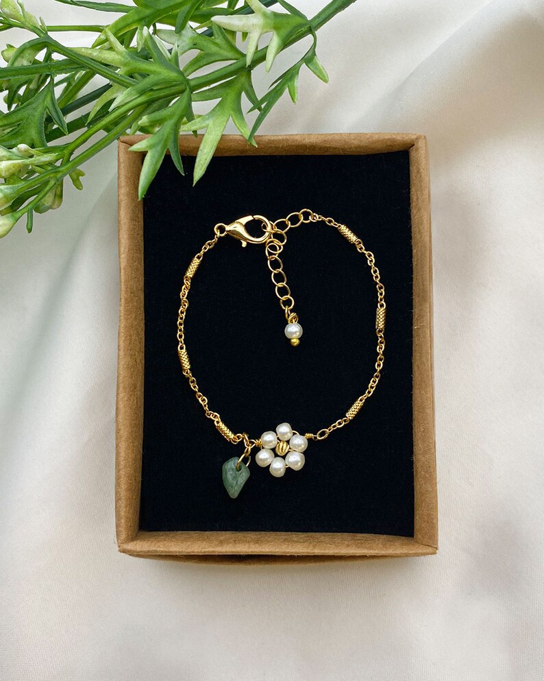 Pu&ograve; includere: Un braccialetto a catena dorata con un delicato ciondolo a forma di fiore realizzato con perle bianche e una pietra verde. Il braccialetto &egrave; esposto in una scatola marrone con un rivestimento in velluto nero.