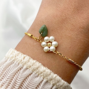 Pu&ograve; includere: Un braccialetto a catena dorata con un ciondolo di pietra verde e un ciondolo a forma di fiore fatto di perle bianche.