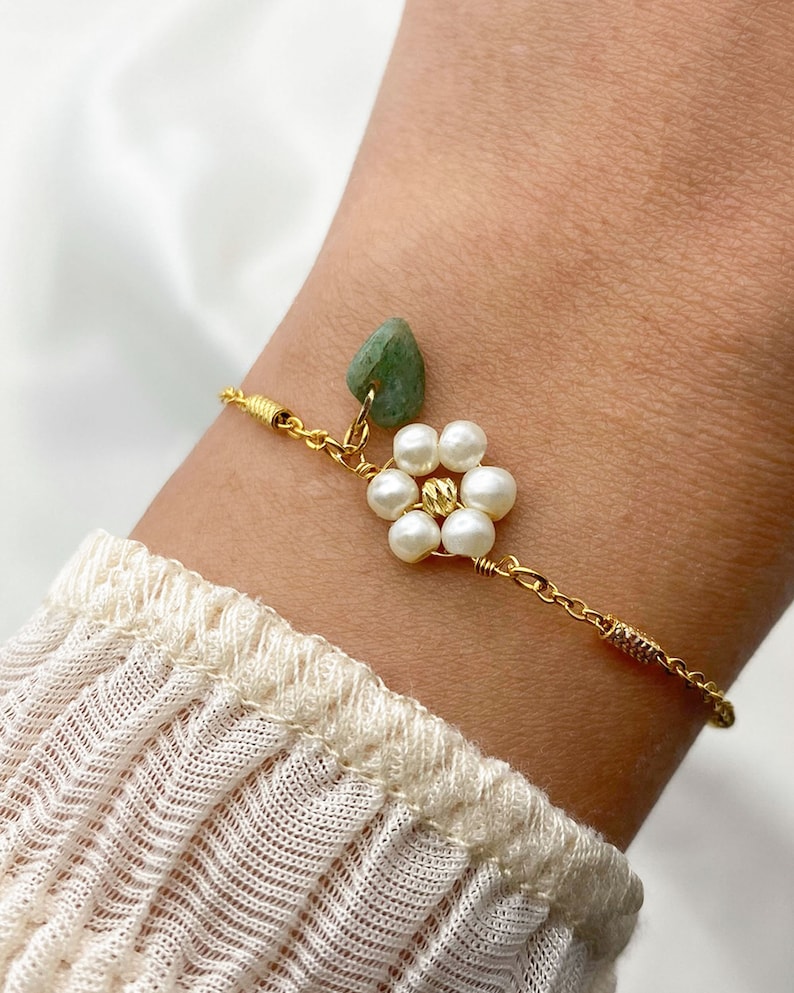 Pu&ograve; includere: Un braccialetto a catena dorata con un ciondolo di pietra verde e un ciondolo a forma di fiore fatto di perle bianche.