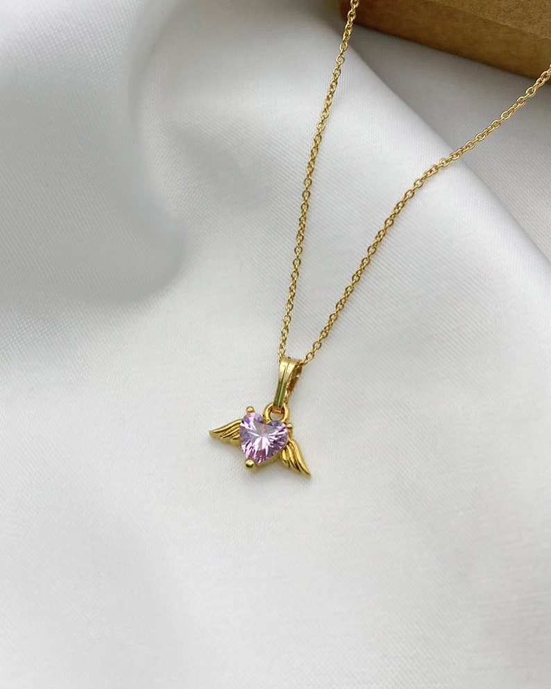 Puede incluir: Un collar de cadena dorada con un colgante de coraz&oacute;n rosa con alas.