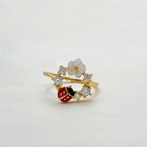 May include: A gold-colored ring featuring a ladybug and flower design. The ladybug is red with black spots and a black head. The flower is white with a yellow center. Clear, round gemstones are set around the ring's band.