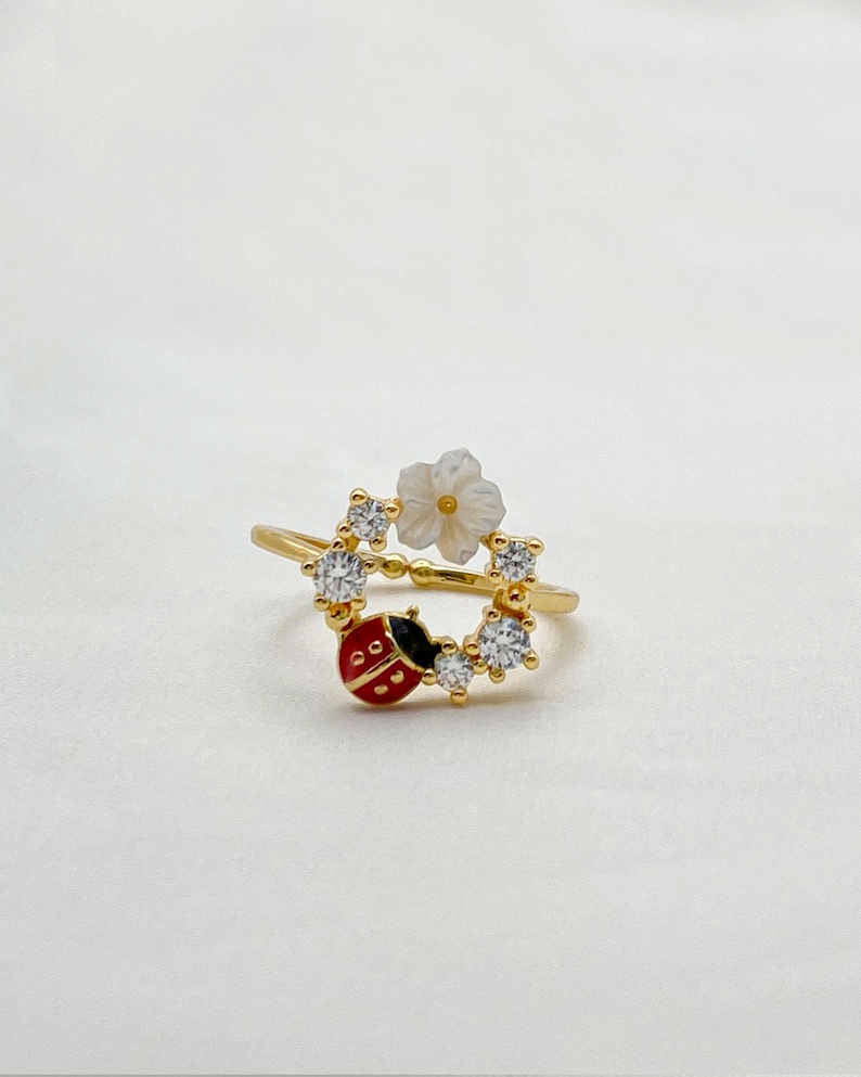 May include: A gold-colored ring featuring a ladybug and flower design. The ladybug is red with black spots and a black head. The flower is white with a yellow center. Clear, round gemstones are set around the ring's band.
