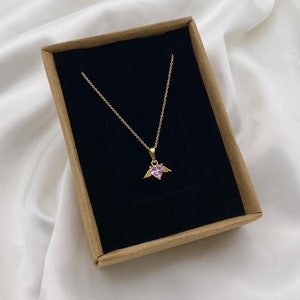 Puede incluir: Un collar de cadena dorada con un colgante de coraz&oacute;n rosa con peque&ntilde;as alas doradas. El collar se muestra en una caja marr&oacute;n con interior negro.