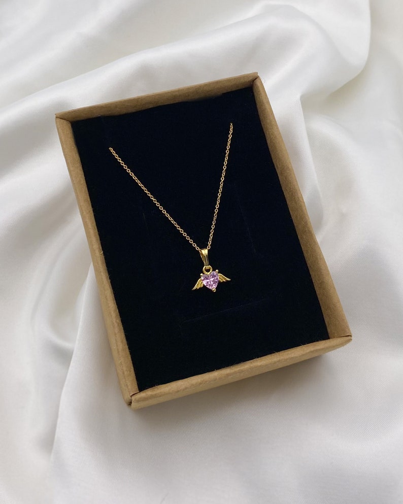 Puede incluir: Un collar de cadena dorada con un colgante de coraz&oacute;n rosa con peque&ntilde;as alas doradas. El collar se muestra en una caja marr&oacute;n con interior negro.