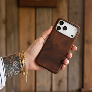 May include: A brown leather phone case with a white camera section. The case is held in a hand with gold bracelets and a ring. The background is a wooden wall with a clock.