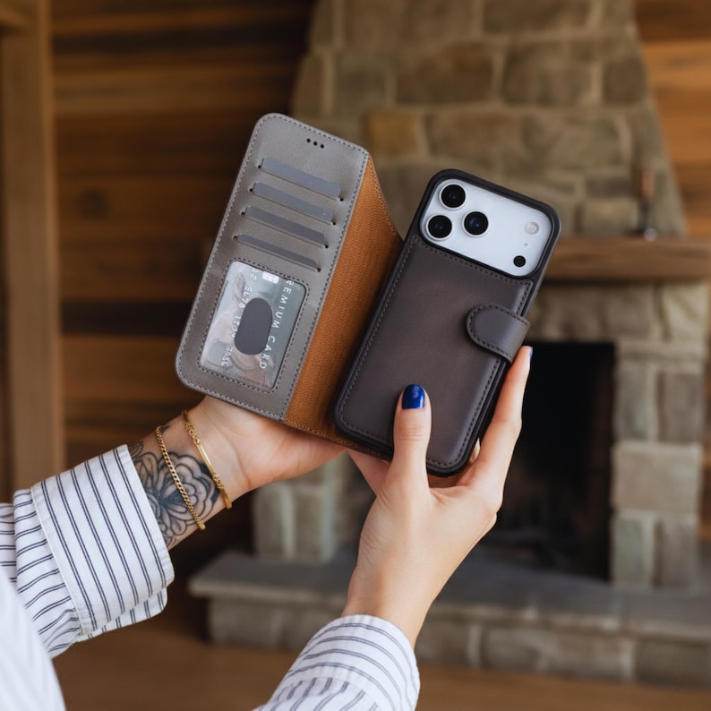 May include: A brown leather phone case with a wallet feature, held open to display the card slots and a credit card. The phone case is open, revealing a gray interior and a brown exterior. The phone case is designed to fit a smartphone.