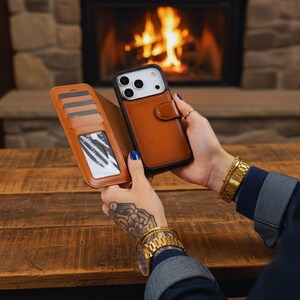 May include: A brown leather phone case with a wallet design, featuring card slots and a snap closure. The phone case is held open, revealing the phone and the wallet section. The background includes a fireplace and a wooden table.