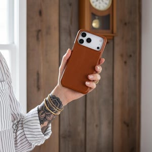 May include: A brown leather phone case with a white camera module. The case is held in a hand with gold bracelets and a floral tattoo. The background features a wooden wall and a vintage clock.
