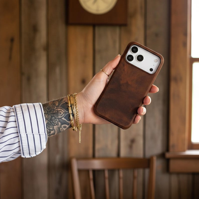 May include: A brown leather phone case with a white camera section. The case is held in a hand with gold bracelets and a ring. The background is a wooden wall with a clock.