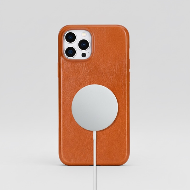 May include: An orange leather phone case with a white circular charger attached. The phone case has a textured surface and a camera cutout. The charger is connected with a white cable.