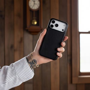 May include: Black phone case with a textured back and a camera cutout. The case is held in a hand, showcasing the design and fit. The background features a wooden wall and a clock.