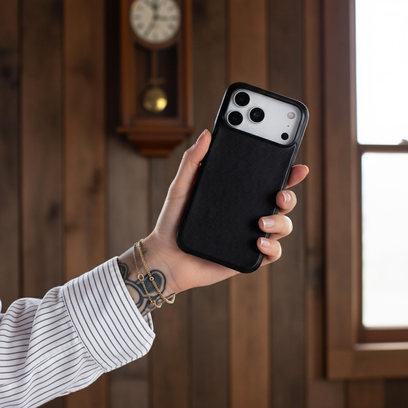 May include: Black phone case with a textured back and a camera cutout. The case is held in a hand, showcasing the design and fit. The background features a wooden wall and a clock.