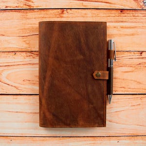 May include: A brown leather journal with a pen tucked into the strap. The journal is lying on a wooden surface.
