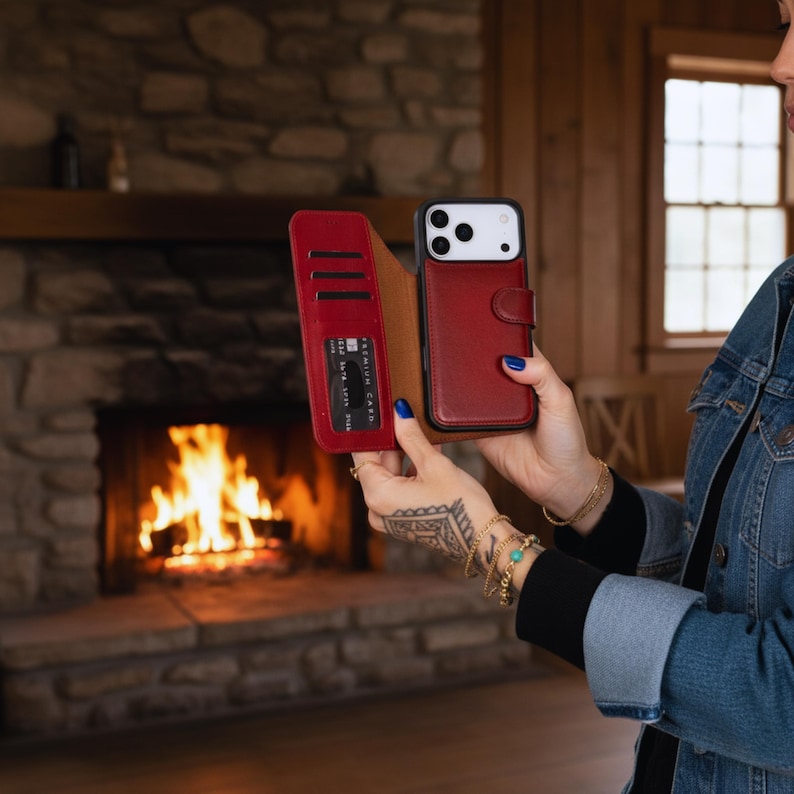 May include: A red leather phone case with a wallet feature, holding a credit card. The case is open, revealing the phone and card slots. The phone has a black frame and white back. The case is held in front of a fireplace.