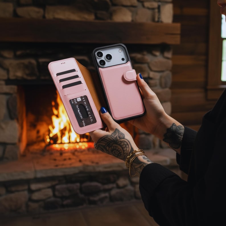 May include: A pink leather phone case with a wallet feature, shown open to reveal card slots and a credit card. The phone case is held next to a closed pink phone case with a black border. The background is a stone fireplace.